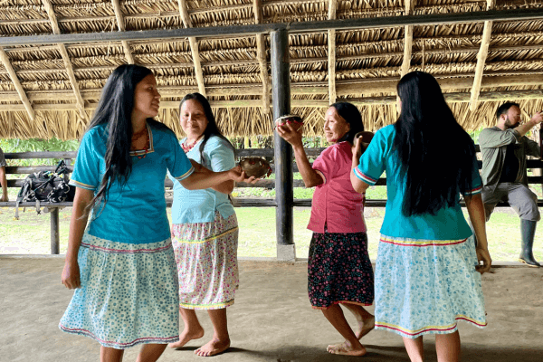 Women from Kichwa community in floral skirts and turquoise tops perform a traditional dance under a thatched-roof pavilion.