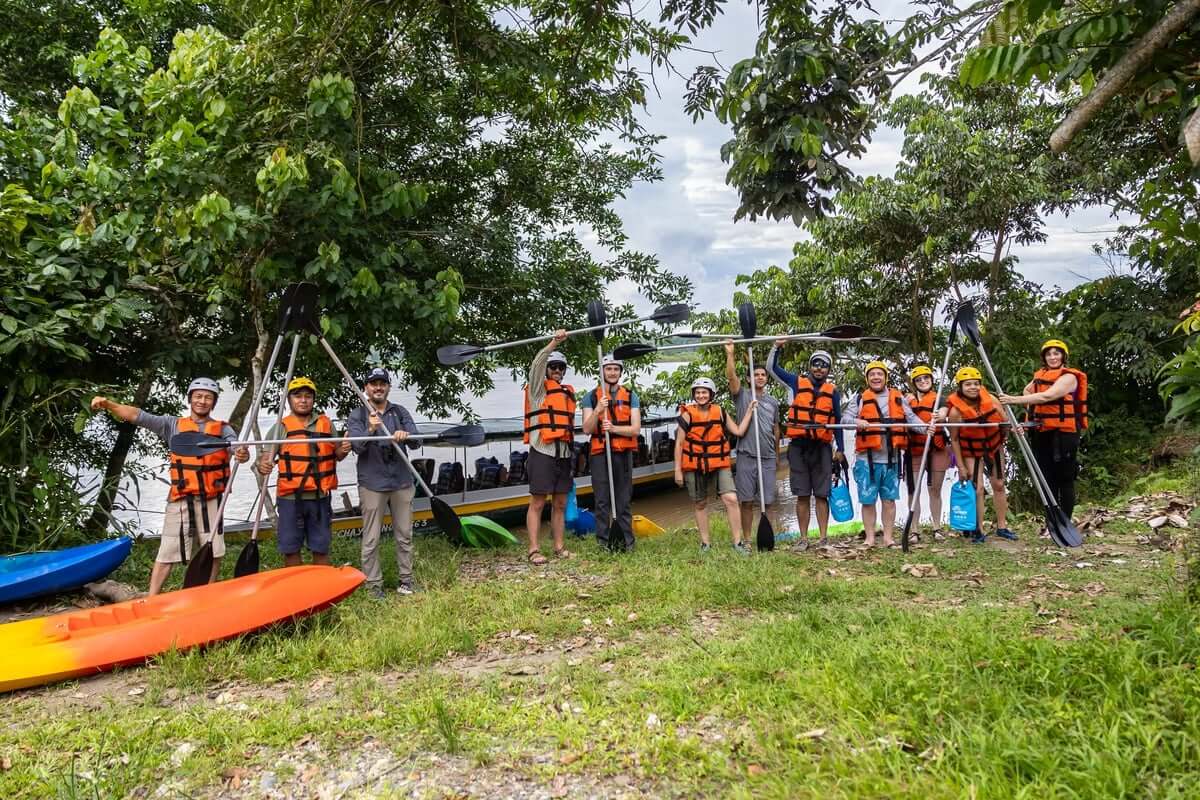 A group of adventure travelers wearing orange life jackets stand along a riverbank, holding kayak paddles upright in an attempt to spell out 'ATTA' (Adventure Travel & Trade Association). Colorful kayaks rest on the grassy shore beside them, and a tour boat is visible in the background. The scene is framed by lush green trees along the Amazonian Napo river.